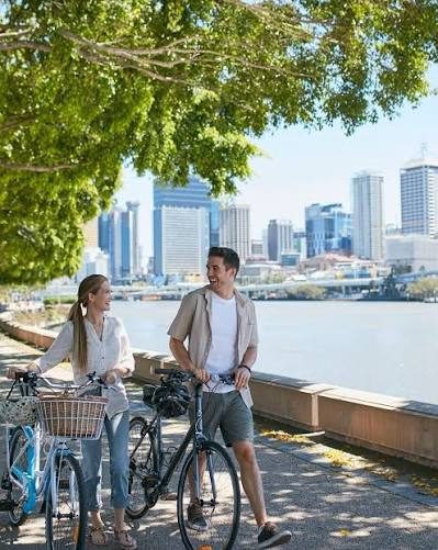 A couple walking city bikes along the Brisbane River with the CBD behind them